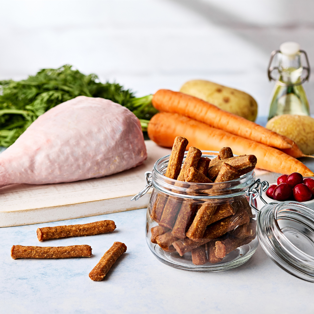 Jar of dog treats on a table with raw meat, carrots, and vegetables in the background.