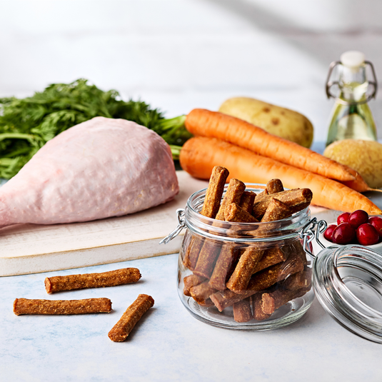 Jar of dog treats on a table with raw meat, carrots, and vegetables in the background.