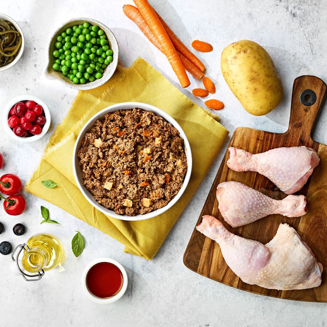 A selection of pet food, alongside visible ingredients such as chicken pieces, peas, carrots, and a potato, arranged on a table with a chopping board and bowls.
