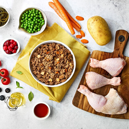 A selection of pet food, alongside visible ingredients such as chicken pieces, peas, carrots, and a potato, arranged on a table with a chopping board and bowls.